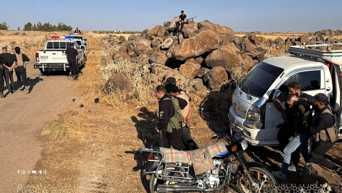 Group of armed men and police near a white van and a motorcycle in a dry, rocky desert area with a large rock pile in the background.