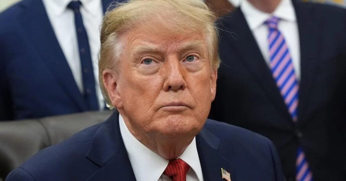 Older man in a navy suit and red tie sits in a formal setting, wearing an American flag lapel pin.