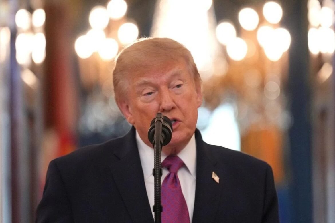 Man in a dark suit and purple tie speaks into a microphone, wearing an American flag pin on the lapel.