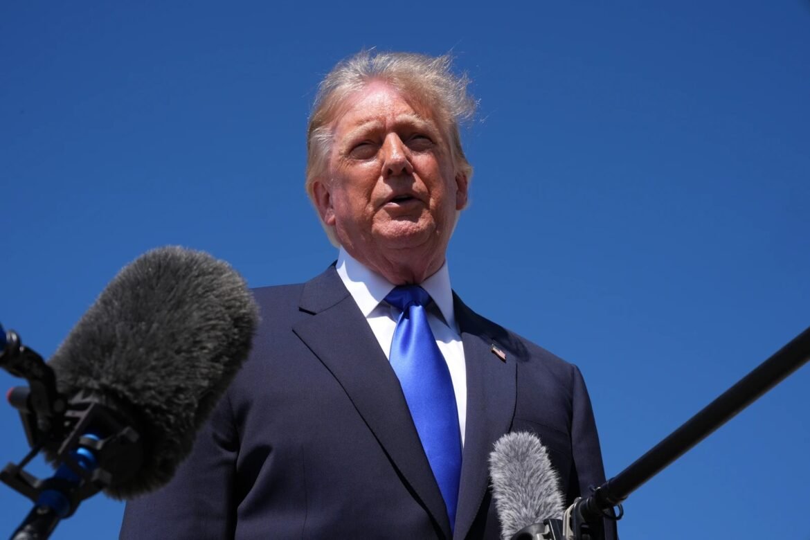 Man in a dark suit and bright blue tie speaks at a press conference outdoors, with several microphones in the foreground and a clear blue sky behind him.