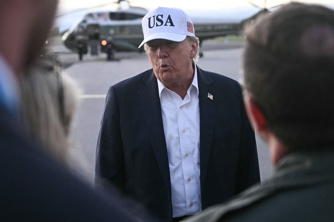 Man in a dark suit and white shirt wearing a white cap with 'USA' speaks to people on a tarmac near a helicopter, outdoors.