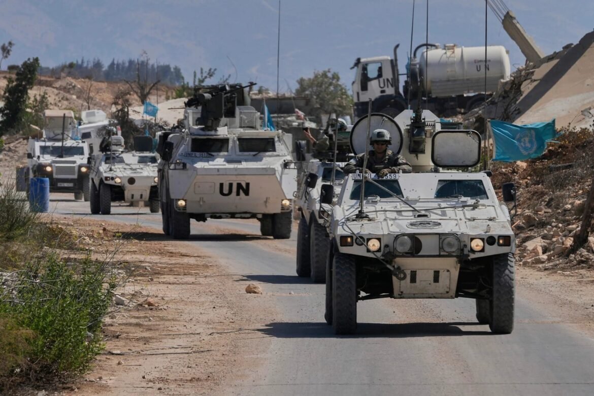 UN armored convoy driving along a dusty desert road, with a blue UN flag visible and rugged hills in the background.