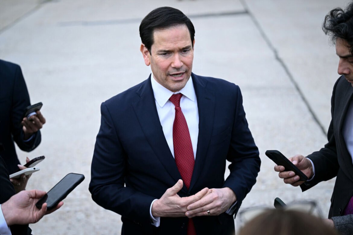 Man in a dark suit with a red tie speaks to reporters holding smartphones outside.