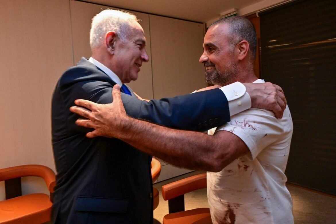 Two men share a warm hug in a hallway; one in a dark suit, the other in a light, stained shirt, both smiling.