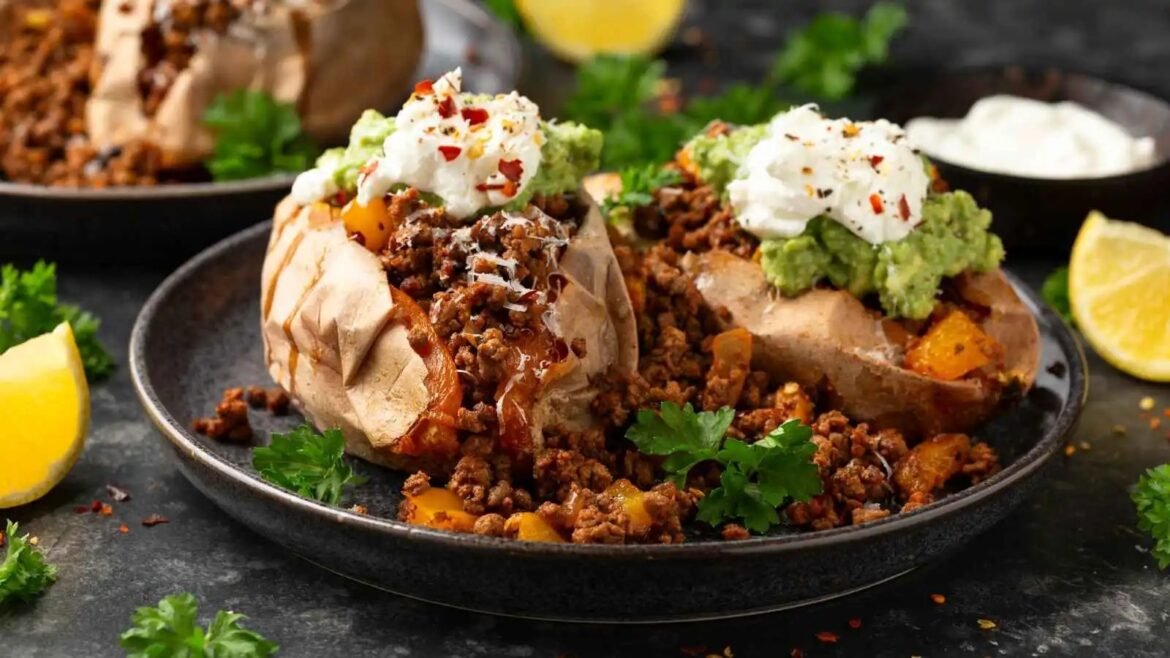 Two loaded baked potatoes on a dark plate, topped with seasoned ground beef, guacamole, sour cream, and red chili flakes, garnished with parsley.