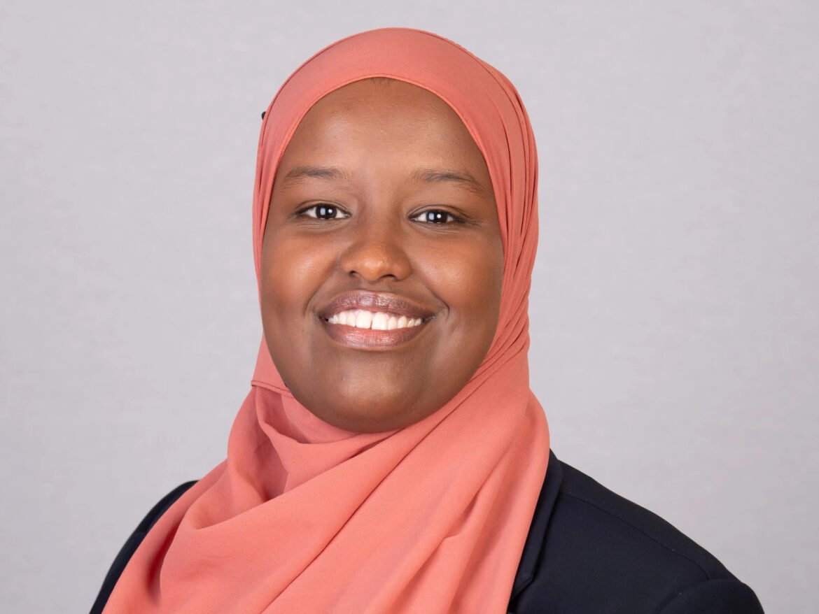 Smiling woman wearing a peach hijab and dark blazer against a neutral gray background, looking at the camera.