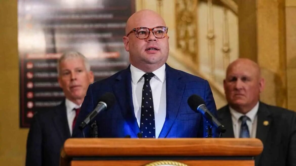 Man in a blue suit and glasses speaks at a wooden podium with two microphones in a formal indoor setting, flanked by two men in suits behind him.