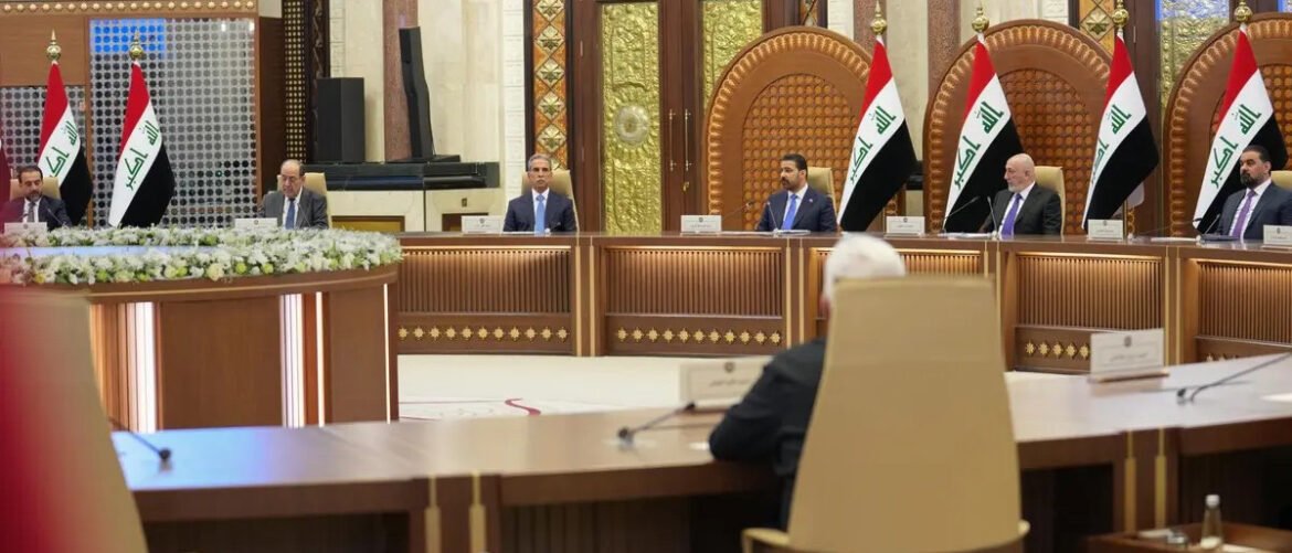 Formal diplomatic meeting in a grand hall with Iraqi flags and officials seated along a curved wooden dais with microphones and nameplates.