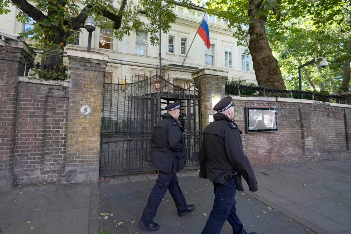 Two police officers in uniform walk past a gated brick wall in front of a white multi-story building with a Russian flag in view.