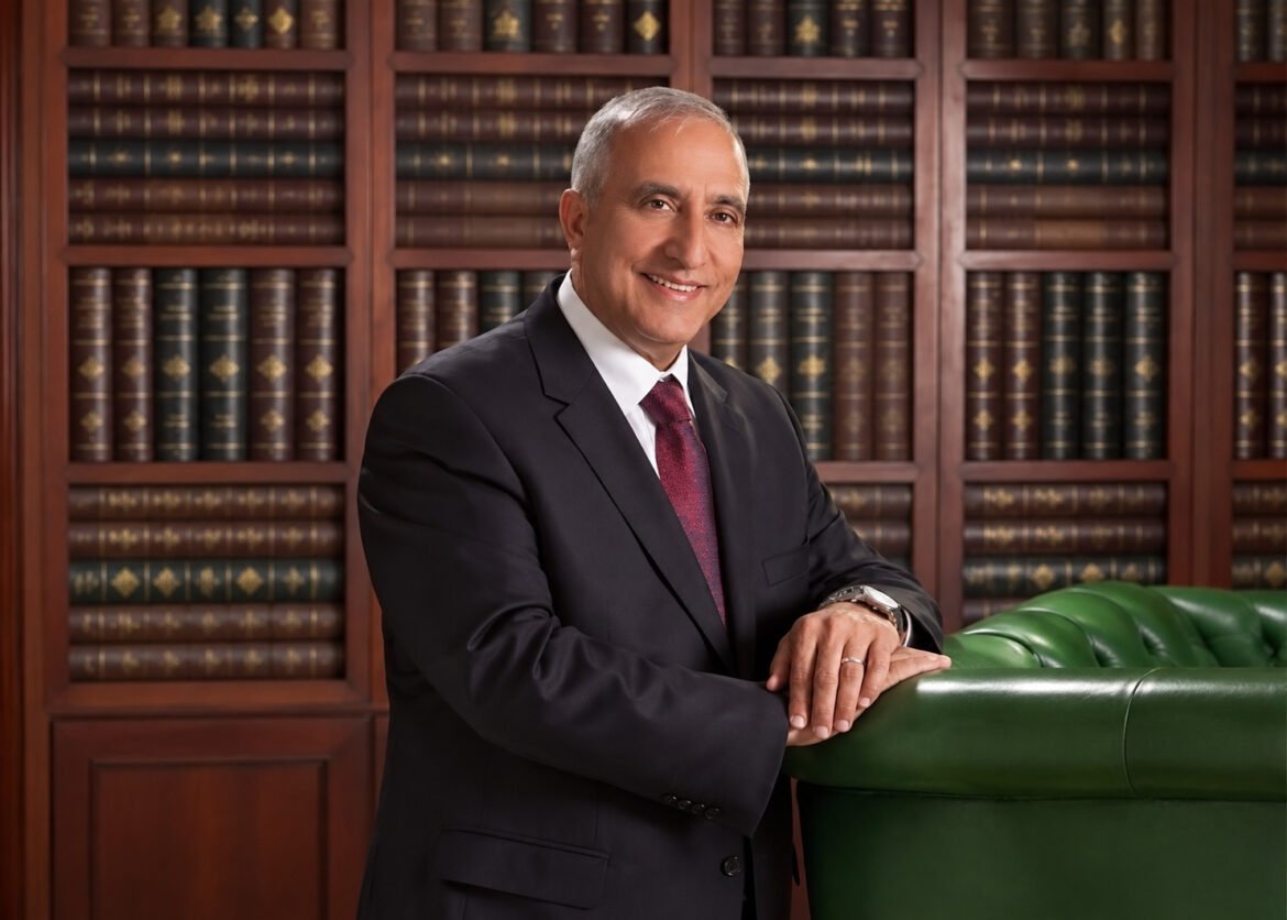 Smiling man in a dark suit and red tie leans on a green leather sofa in front of tall wooden bookcases filled with leather-bound volumes.