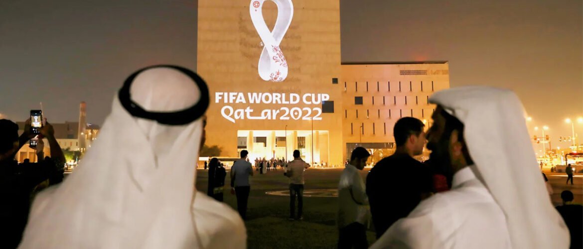 Night scene outside a stadium with a large 'FIFA World Cup Qatar 2022' sign; people in traditional dress and others taking photos.