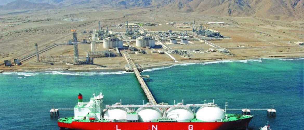 Large LNG carrier with white spherical tanks docked at a pier, approaching an industrial port along a coastal landscape.