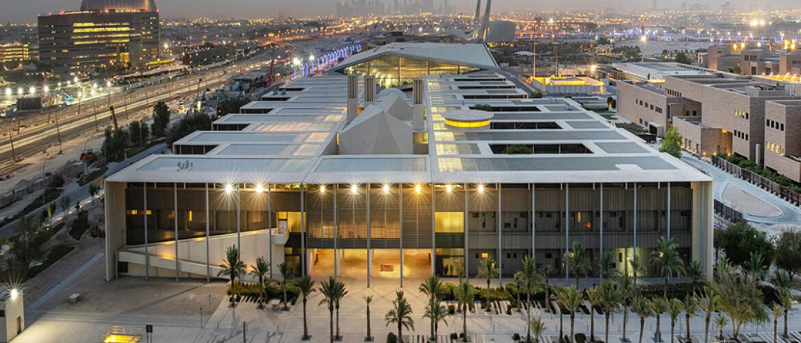 Aerial view of a large modern building with a glass entrance, columns, and palm trees in a plaza at dusk, city lights in the background.