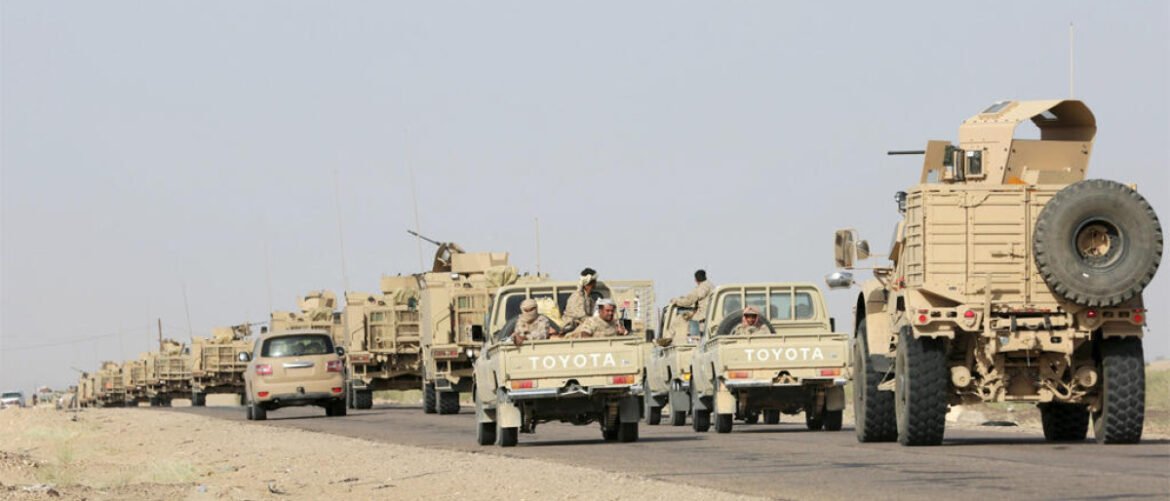 Desert road convoy of beige military vehicles including Toyota pickup trucks and armored cars with soldiers riding in the back bays.