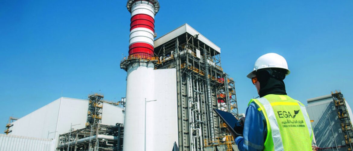 Industrial power plant exterior with a tall white and red striped stack and complex piping, while a worker in a hard hat and high-visibility vest surveys the site with a tablet/clipboard.