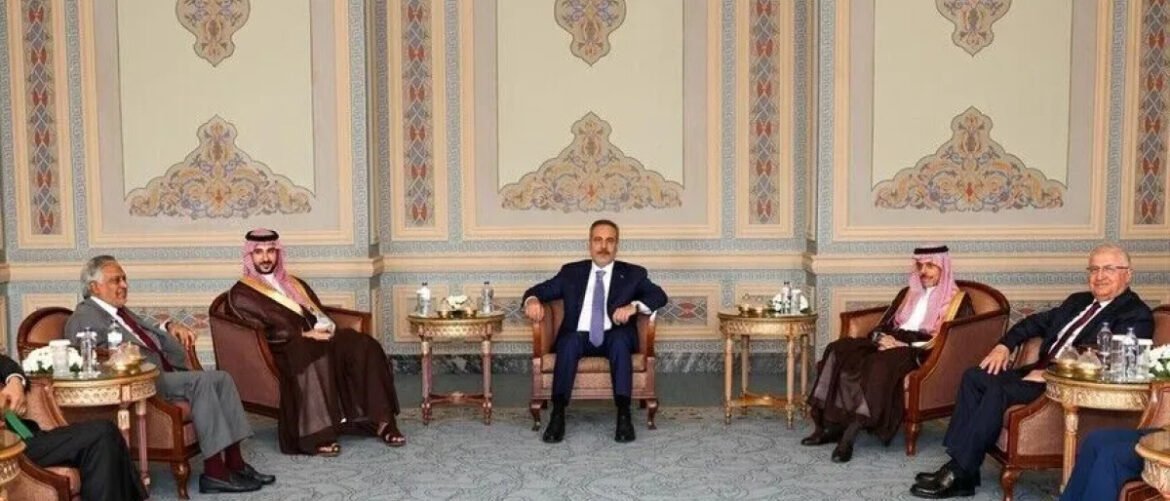 Six men sit in a semicircle of chairs in a richly decorated room, likely a formal meeting.