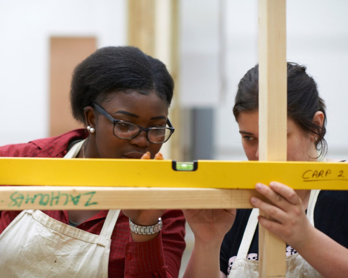 Two women in aprons level a wooden frame using a yellow bubble level in a workshop setting.