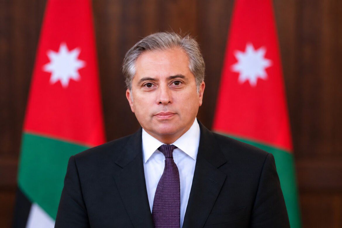 Professional portrait of a middle-aged man in a dark suit and purple tie, with Jordanian flags in the background.