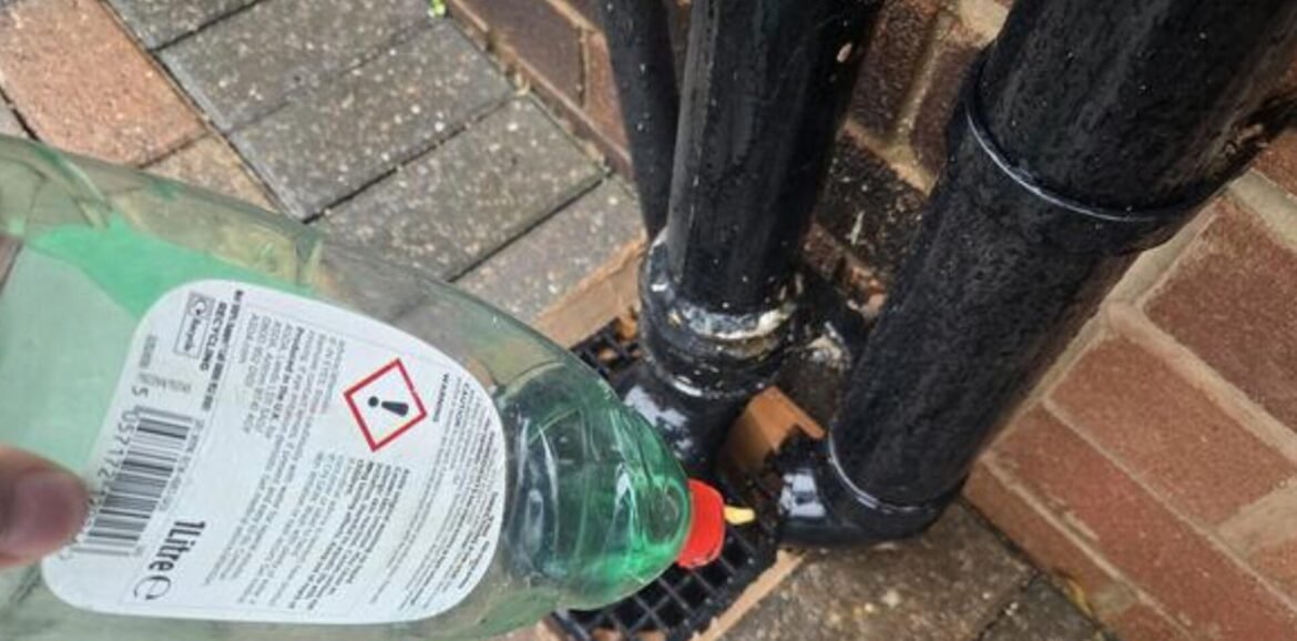Green plastic cleaning bottle with a red-cap and warning label containing a hazard exclamation symbol, placed next to black pipes on a brick surface