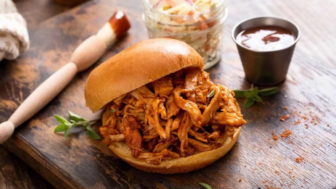 Pulled pork sandwich on a soft bun with tangy barbecue meat spilling out, on a rustic wooden board with a cup of sauce nearby