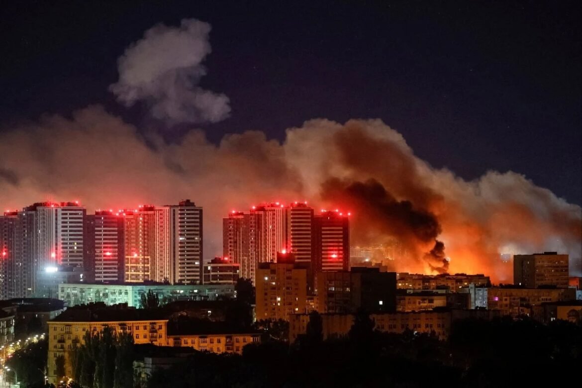 Nighttime city skyline with tall residential buildings illuminated by red lights as a large fire burns in the distance, producing thick orange smoke.