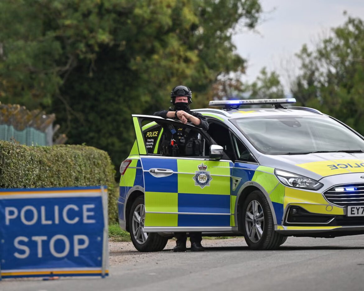 Officer standing by an open car door of a marked police vehicle with blue lights, blocking a road.