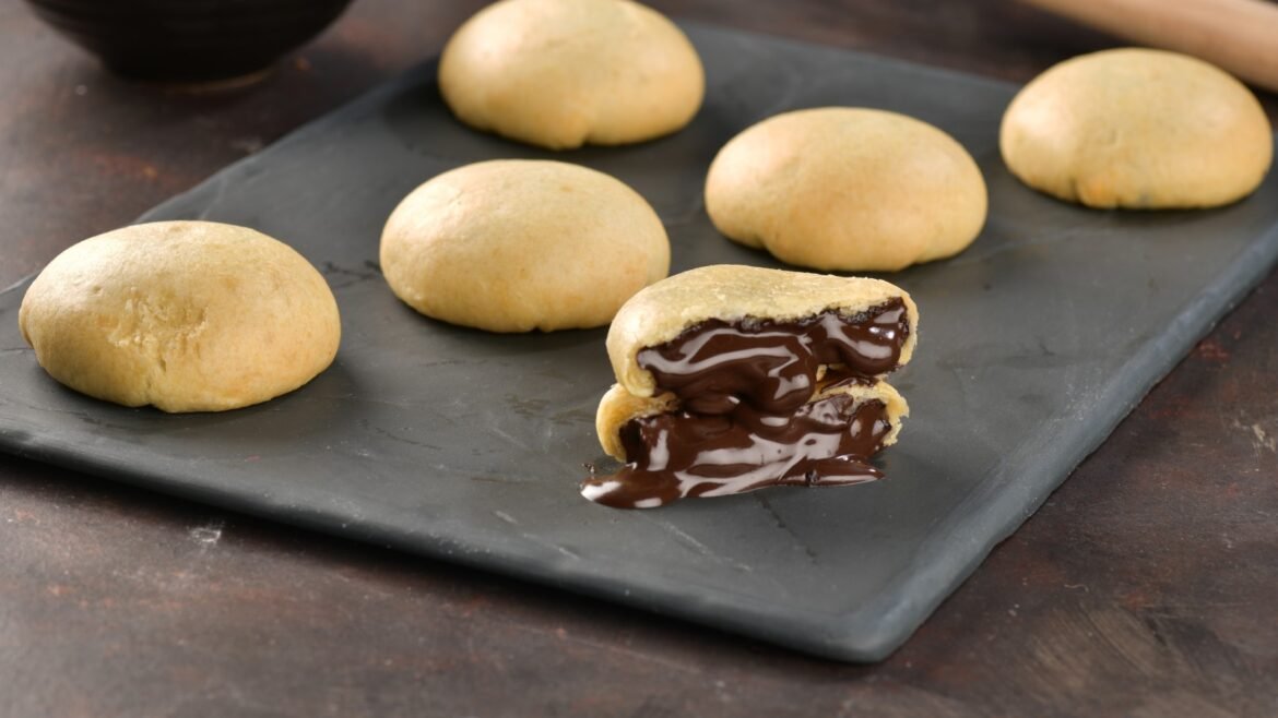 Six round golden cookies on a dark slate tray; one cookie is broken open, revealing a gooey melted chocolate center.