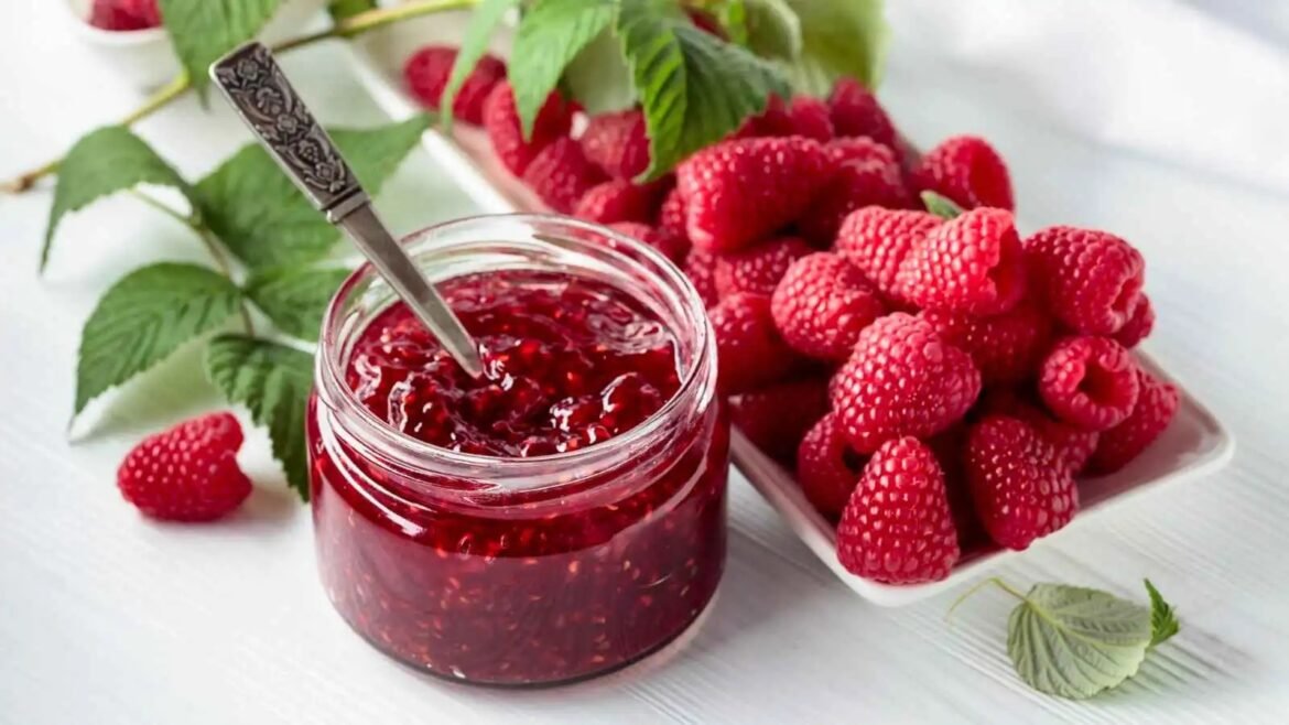 Jar of raspberry jam with a knife, surrounded by fresh raspberries on a white wooden surface.