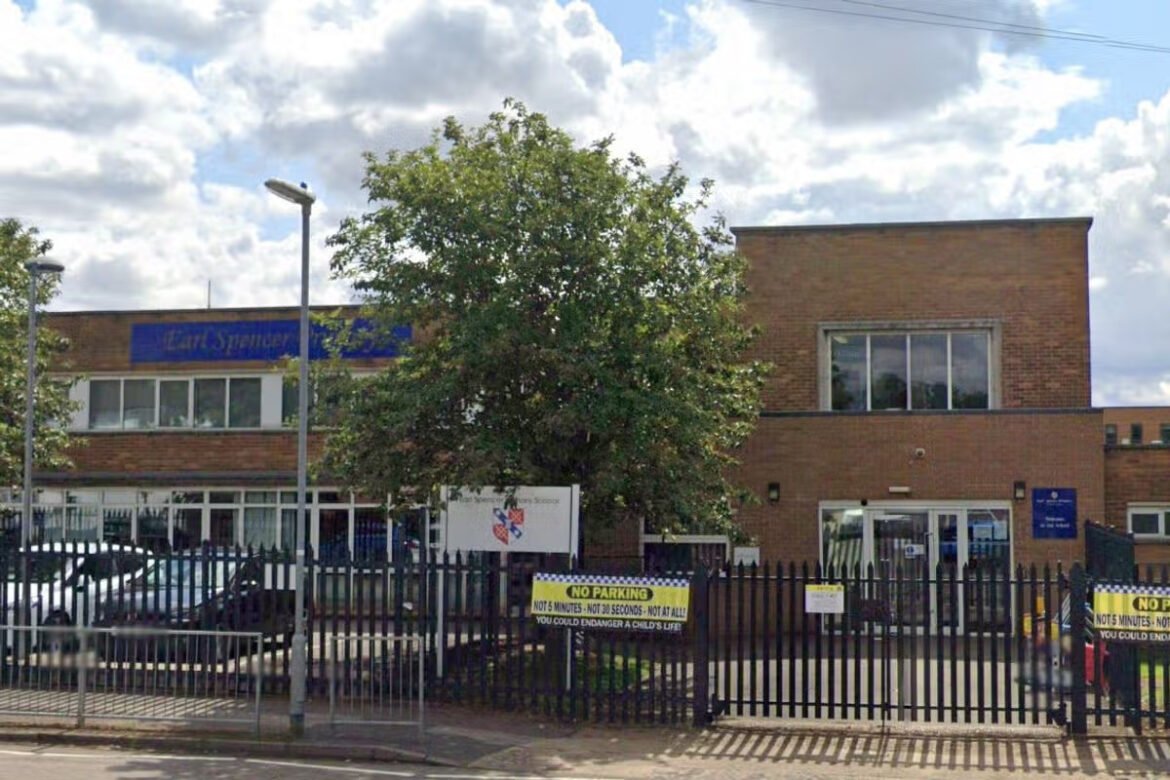 Brick school building behind a black metal fence, with a large tree in front and 'No Parking' signs along the gate area.