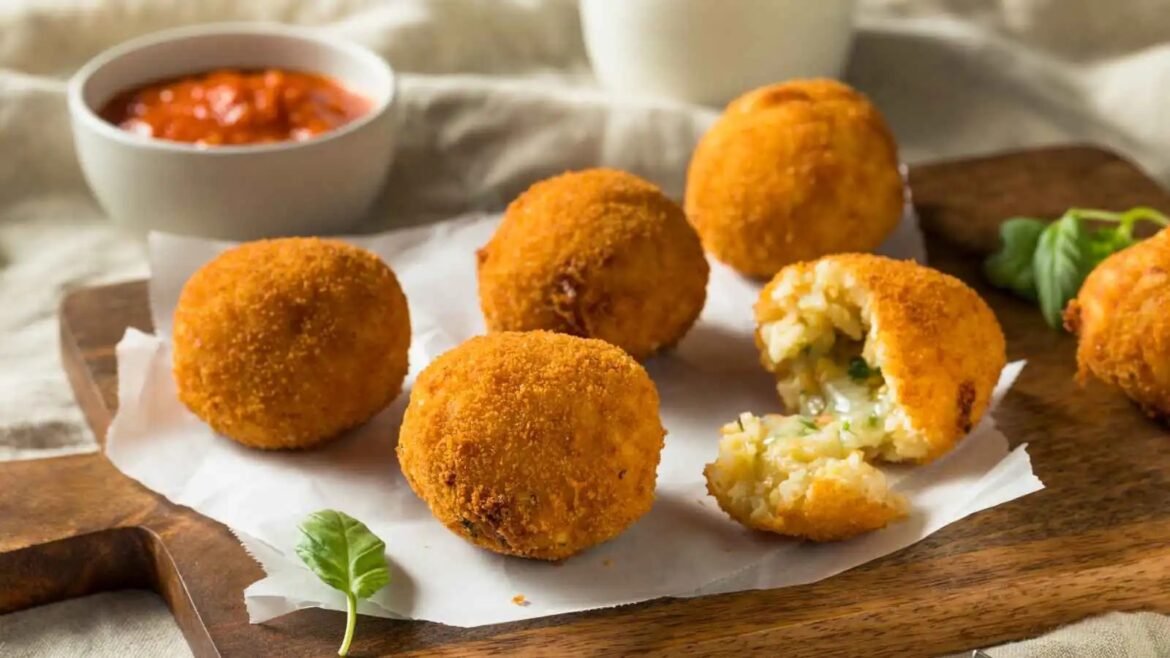 Golden fried croquettes on parchment on a wooden board, with a bowl of marinara sauce in the background; one is broken open to reveal a cheesy potato filling.