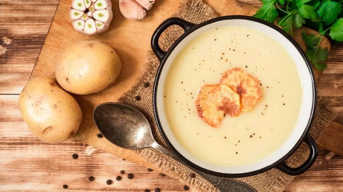 Creamy potato soup in a white pot with black rim, topped with croutons, on a wooden table with potatoes, garlic, and parsley nearby