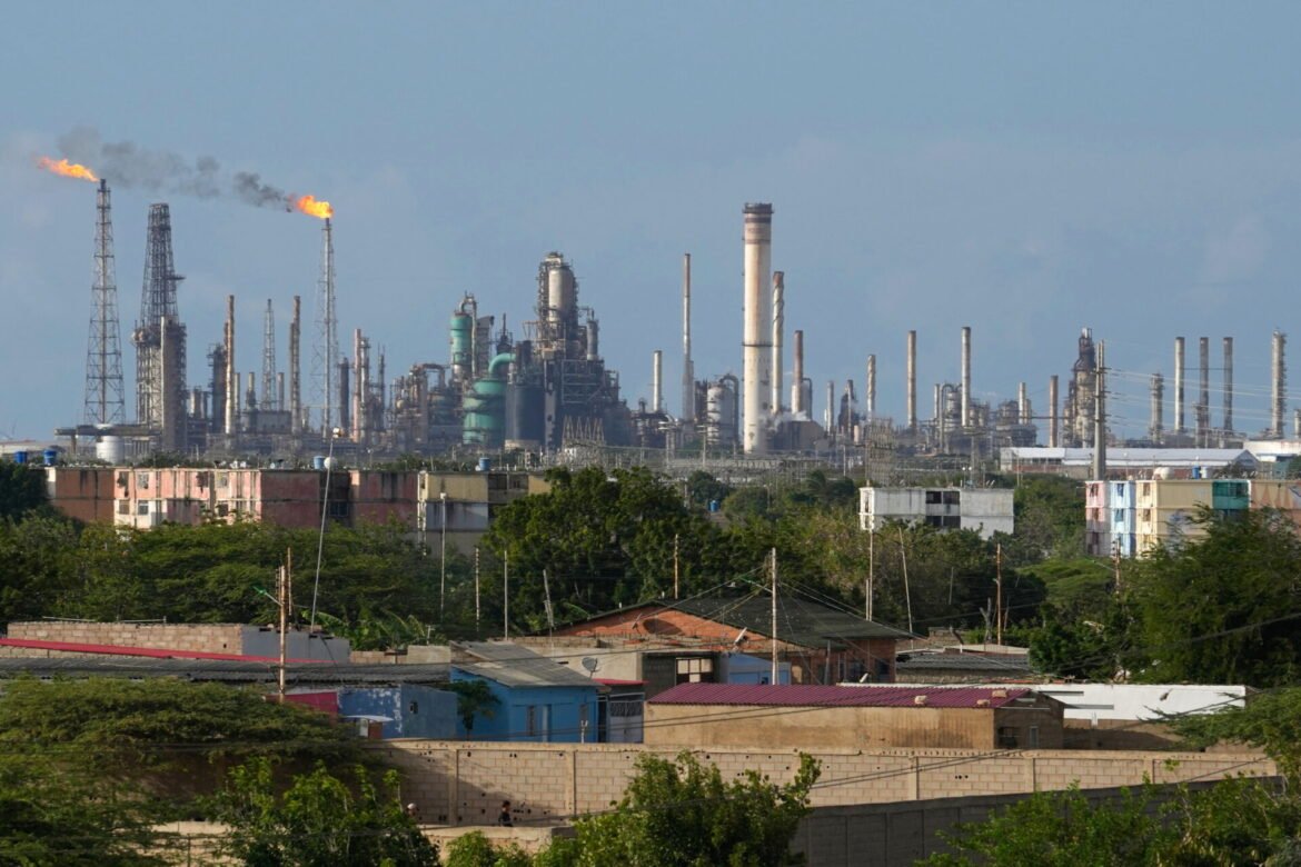 Industrial refinery skyline with numerous smokestacks, towers, and a distant flame plume against a blue sky.