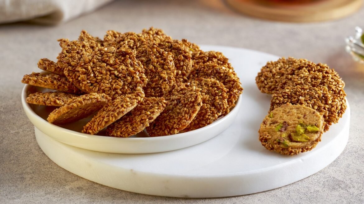Plate of sesame-coated cookies with pistachio-filled centers, some broken into slices on a white dish and cutting board surface.