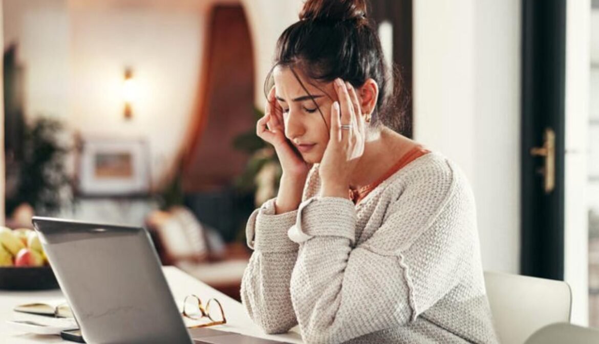 Woman in a cozy sweater holds her temples in frustration while working on a laptop at a kitchen table.