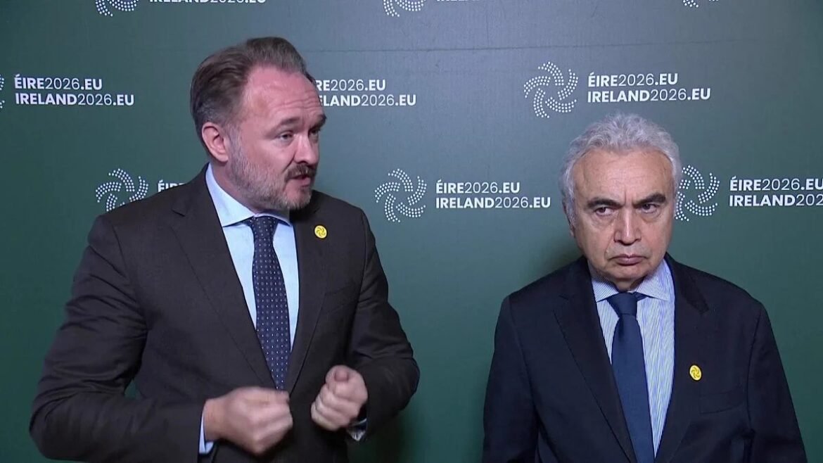 Two men in dark suits stand in front of a green backdrop with ÉIRE2026.EU / IRELAND2026.EU logos; one is gesturing with his hands.
