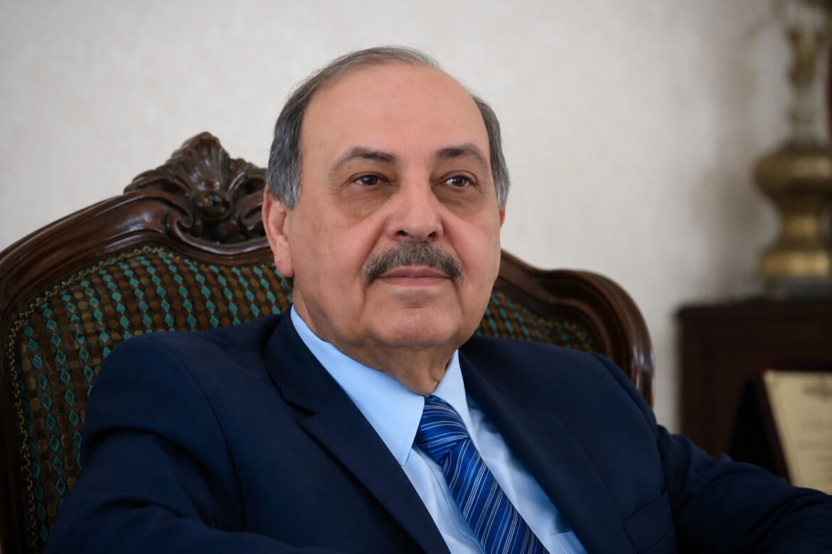 Older man in a blue suit and striped tie sits in an ornate carved chair, looking slightly to the left.
