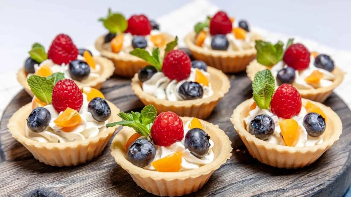 Mini fruit tarts with whipped cream, raspberries, blueberries, mango cubes, and mint on a wooden board.