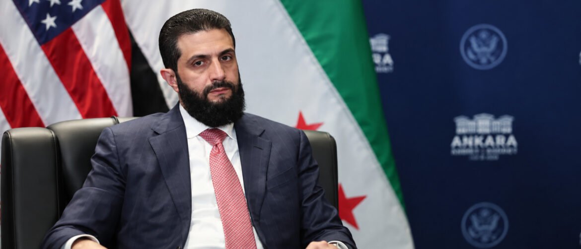 Bearded man in a dark pinstripe suit and pink tie seated at a conference, with US and other flags behind him.