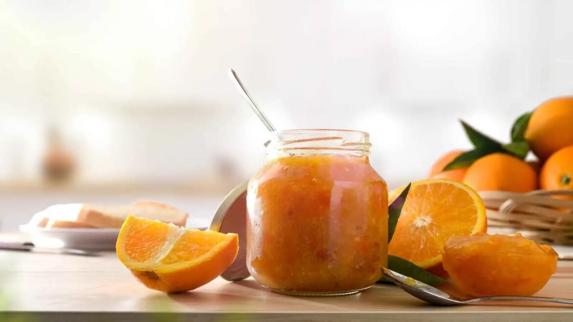 Jar of orange marmalade with a spoon on a light kitchen table, surrounded by orange slices and a basket of oranges in the background.