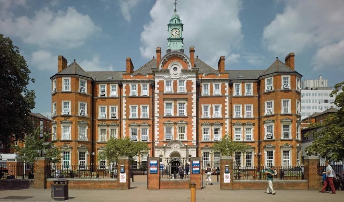 Historic red-brick town hall with a central clock tower, white stone detailing, and symmetrical wings; gate and people outside