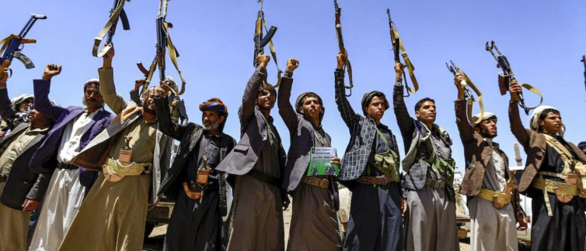 Line of men in traditional clothing raising assault rifles in the air against a clear blue sky, appearing to show a show of force or solidarity.