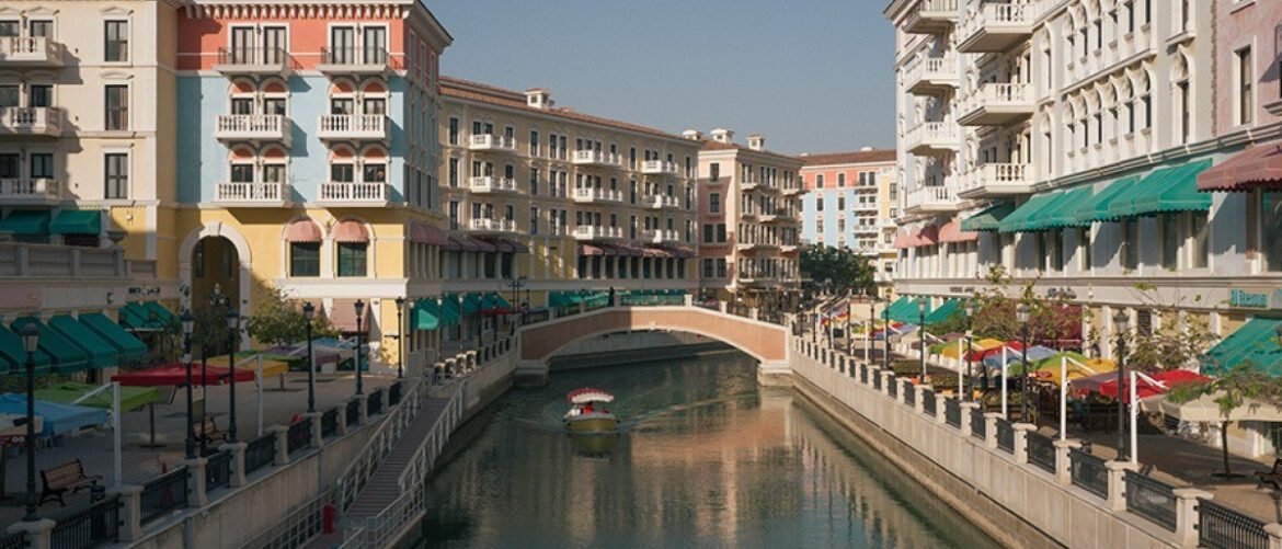 Colorful riverfront street with arched balconies, canal, and a small boat passing under a stone bridge in a sunny town.