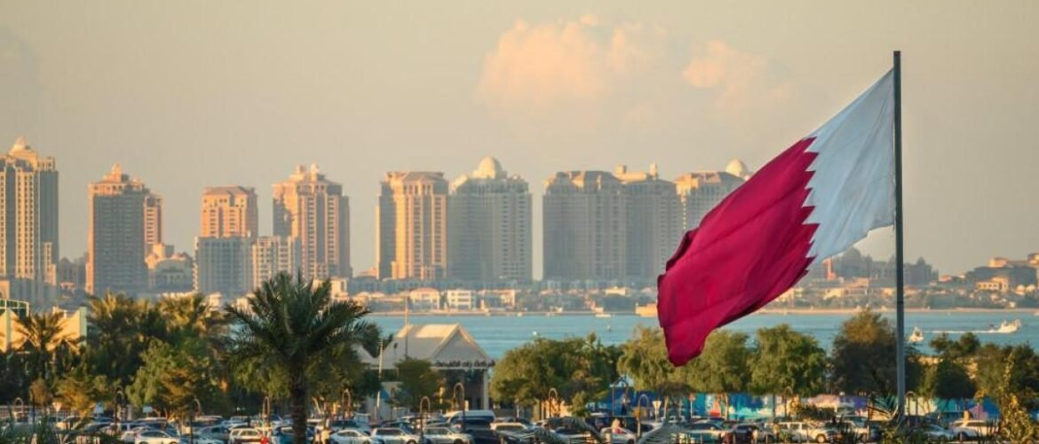 Qatar flag waving in the foreground with a city skyline and palm trees along the coast in Doha, including a busy parking lot.