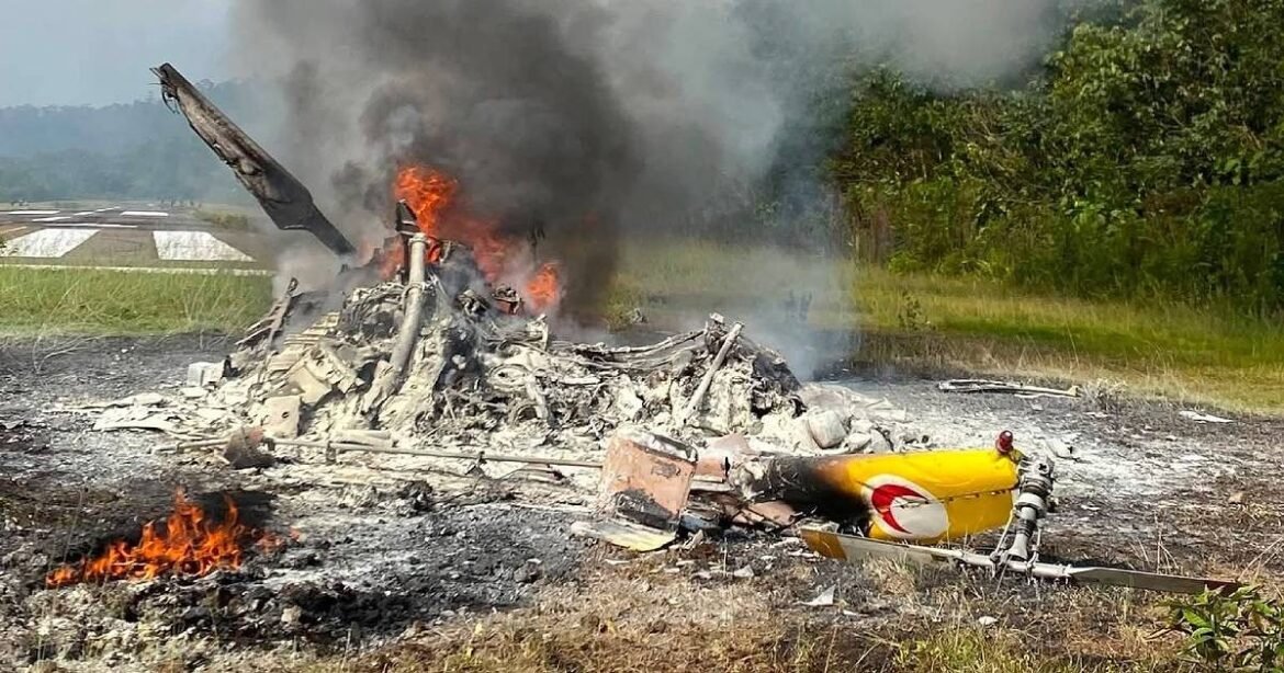 Smoldering aircraft wreckage in a field with flames and thick black smoke rising from the collapsed fuselage and scattered debris nearby.