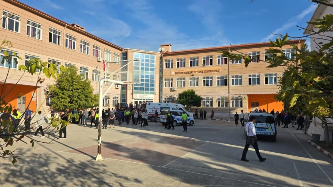 People gather in the school courtyard in front of a large, orange-brick, multi-story building with ambulances and a police car on the scene during daytime.