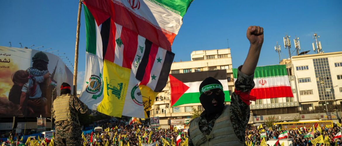 Masked protester raising a clenched fist among a crowd with many flags (Iran, Palestine, and others) in a city square during a rally behind a soldier guard person nearby.