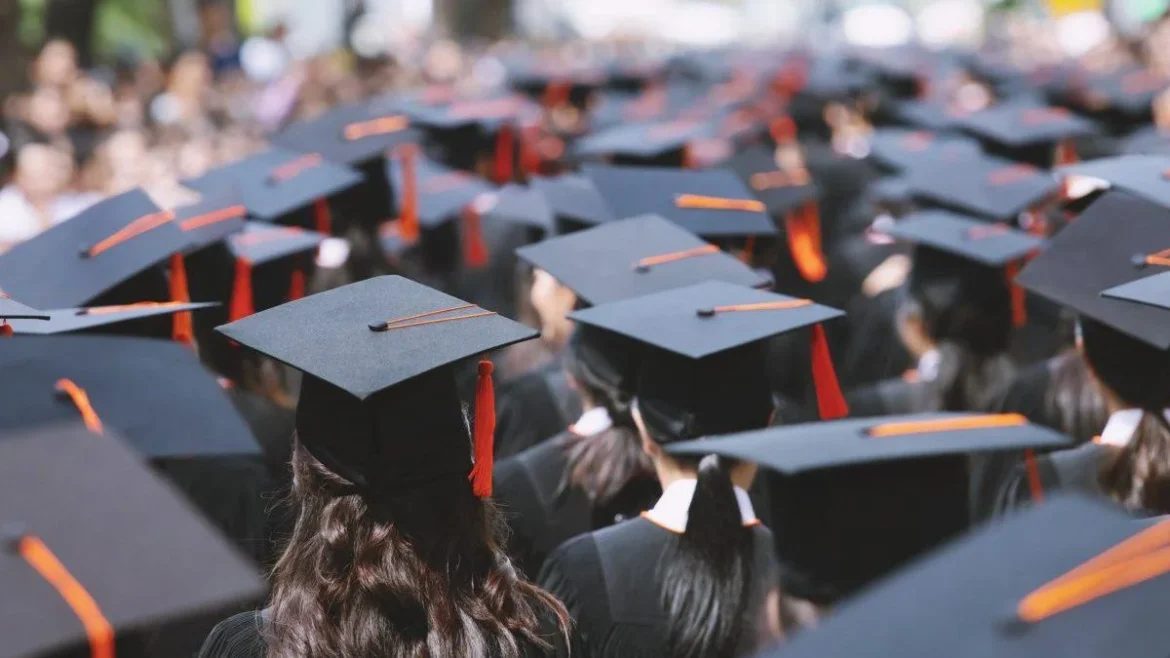 Graduates in black caps and gowns, viewed from behind at a commencement ceremony with orange tassels running across the crowd.