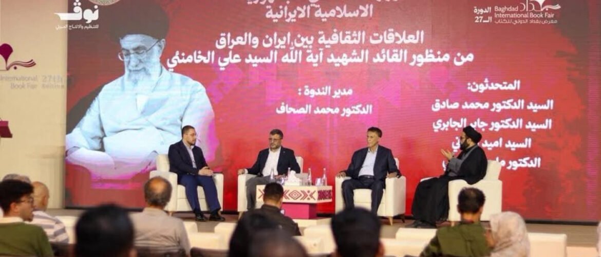 Panel discussion on a stage with four men in suits seated in white chairs, audience in front, a red backdrop with Arabic text and a large portrait image behind them.