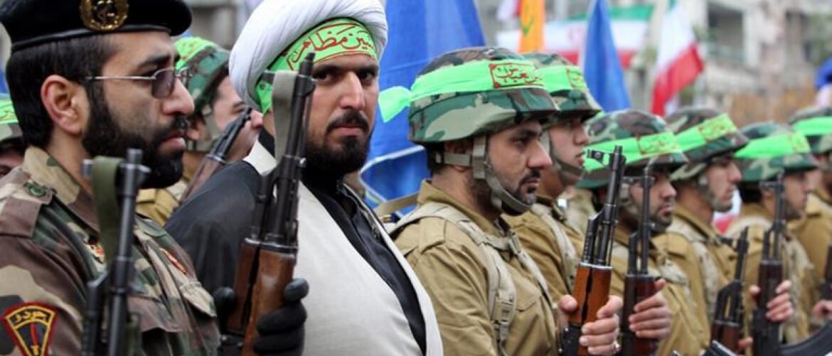 Line of armed soldiers in desert camouflage standing in formation, holding rifles with green headbands visible on their helmets or heads.