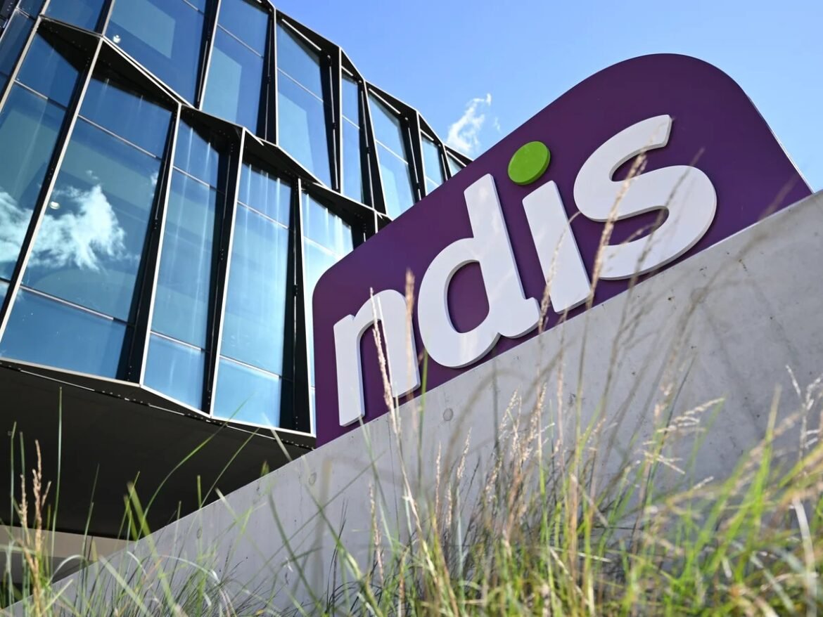 Modern glass-office building with a large purple sign displaying the white letters 'ndis' and a green dot, set against a blue sky.